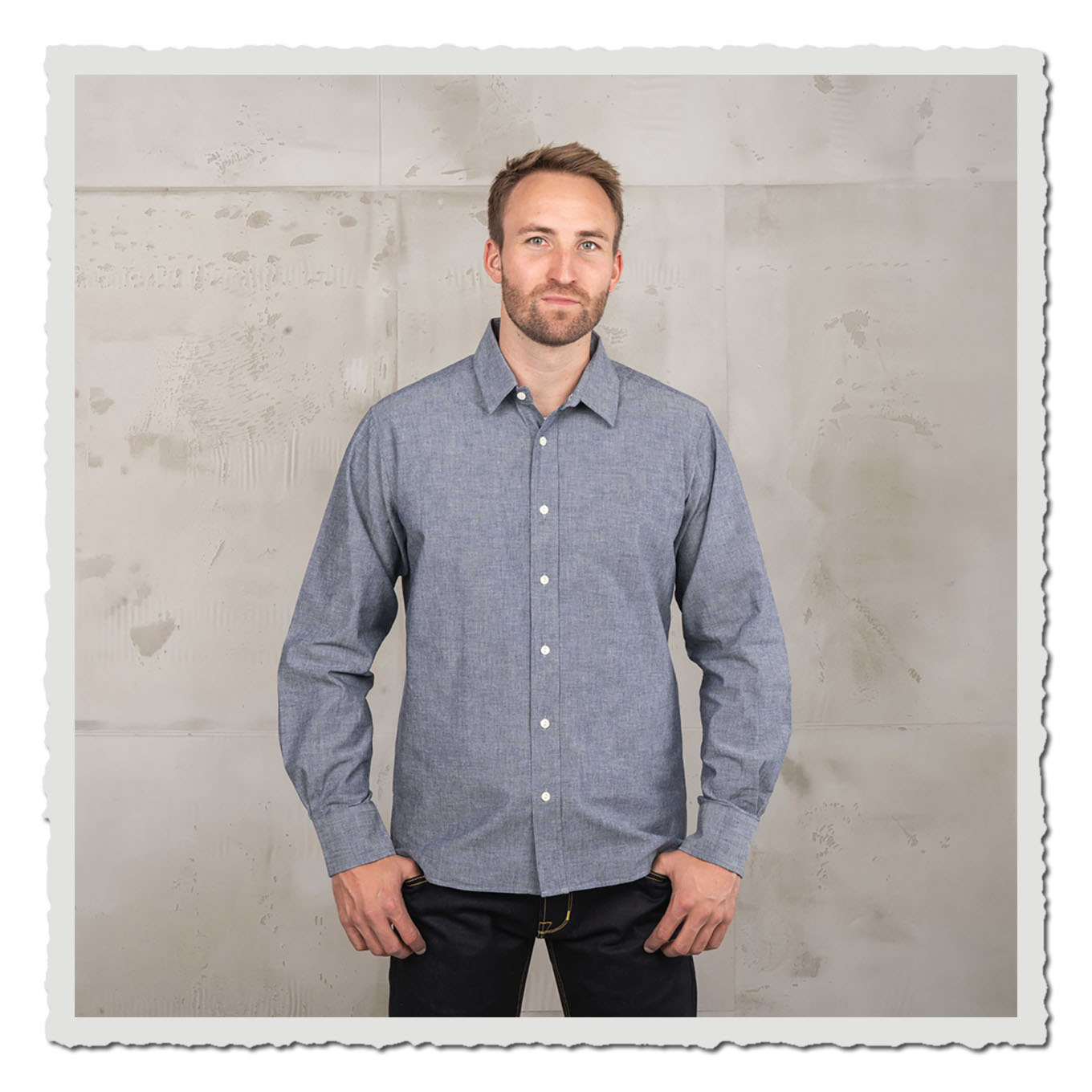 Person mit blauem Langarmhemd und dunkler Jeans vor hellem, neutralem Hintergrund im Studio.